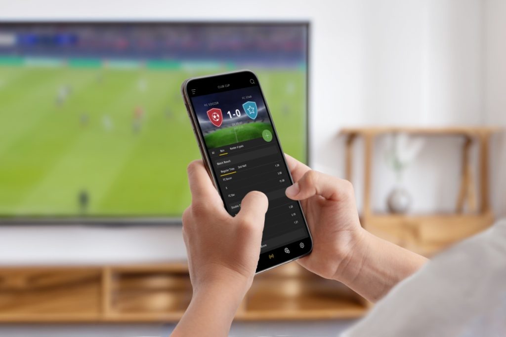 barbat tinand in mana un telefon mobil pe care este afisat scorul si cotele unui meci