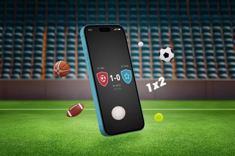 telefon mobil afisand rezultatul live al unui meci de fotbal, cu un teren in fundal