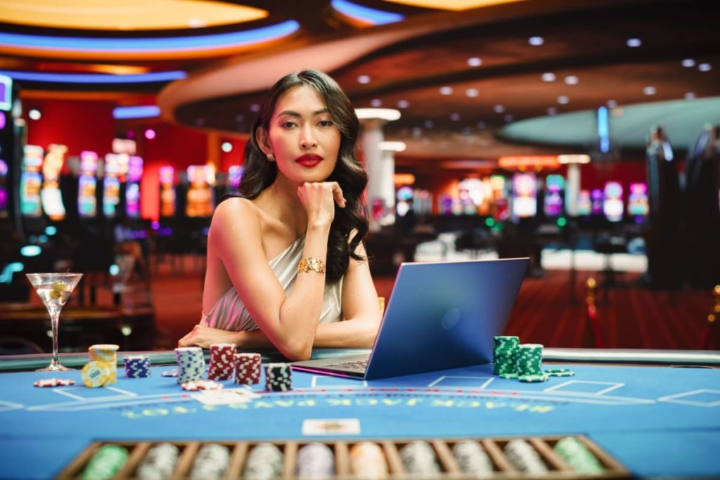 femeie crupier la o masa de baccarat live online in fata unui laptop cu jetoane de cazino in jur