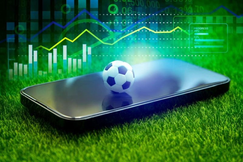 telefon mobil si minge de fotbal pe gazonul unui teren de fotbal cu statistici in fundal