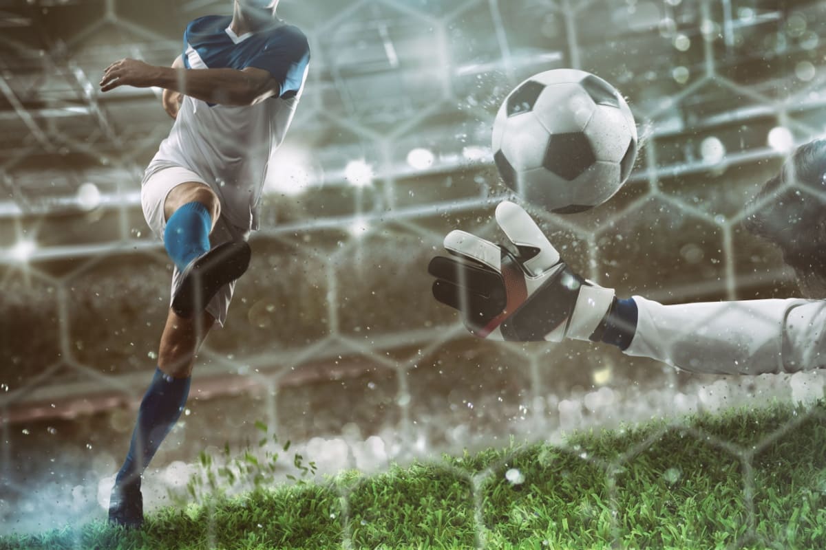 jucator sutand la poarta in timpul unui meci de fotbal, in timp ce portarul advers incearca sa apere