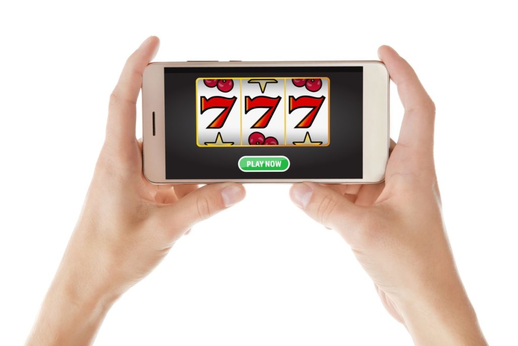 doua maini tinand un telefon pe care ruleaza un joc de slot cu fructe si septari