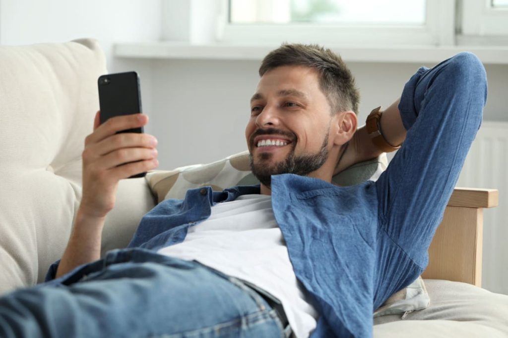 barbat zambitor intins pe canapea, cu un telefon mobil in mana