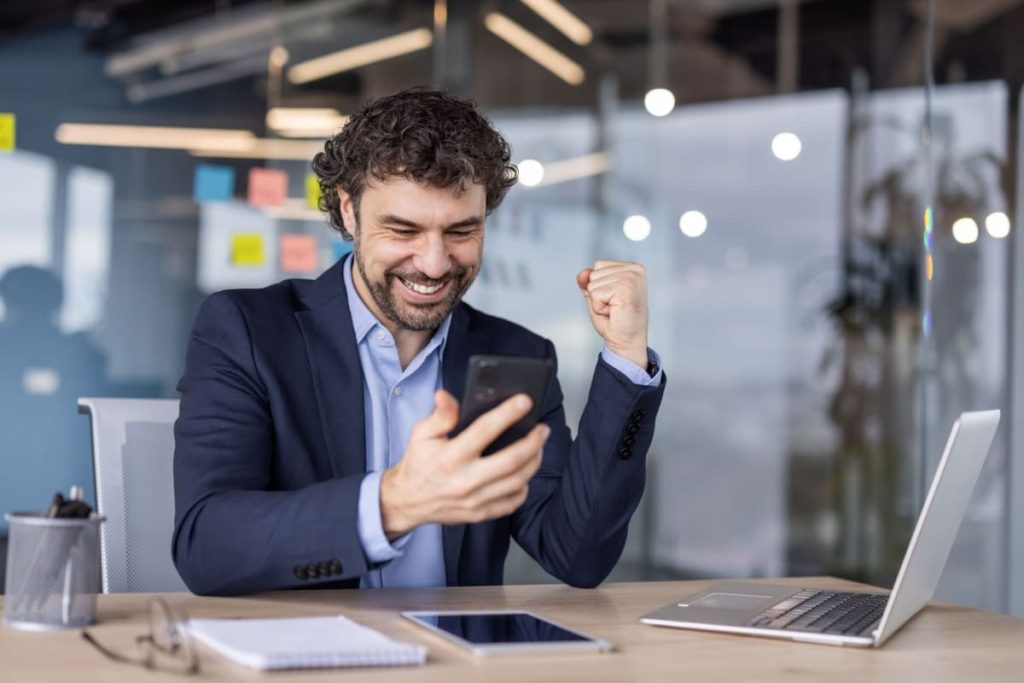 barbat fericit, asezat la birou, tinand in mana un telefon mobil