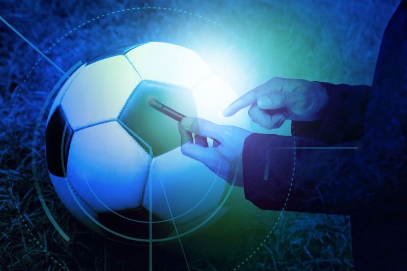 barbat tinand in mana un telefon mobil; o minge de fotbal pe teren in fundal