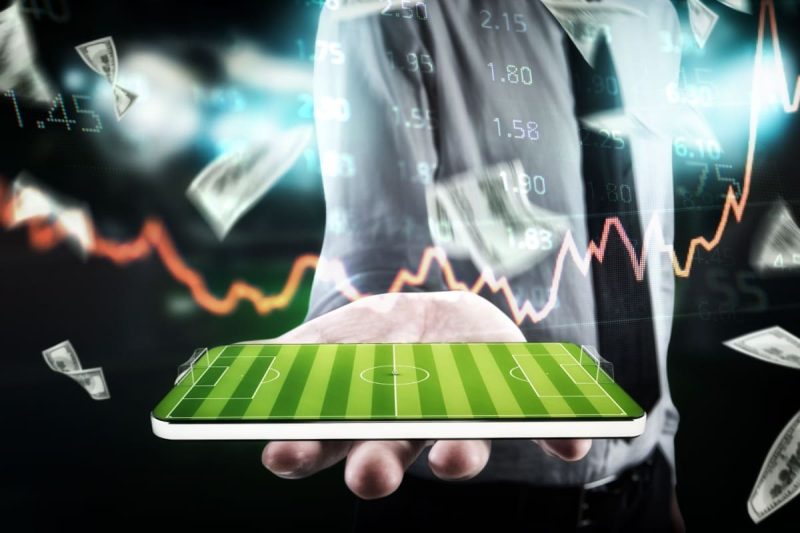barbat tinand in mana un telefon mobil care afiseaza un teren de fotbal, cu grafice, cote si bancnote in fundal