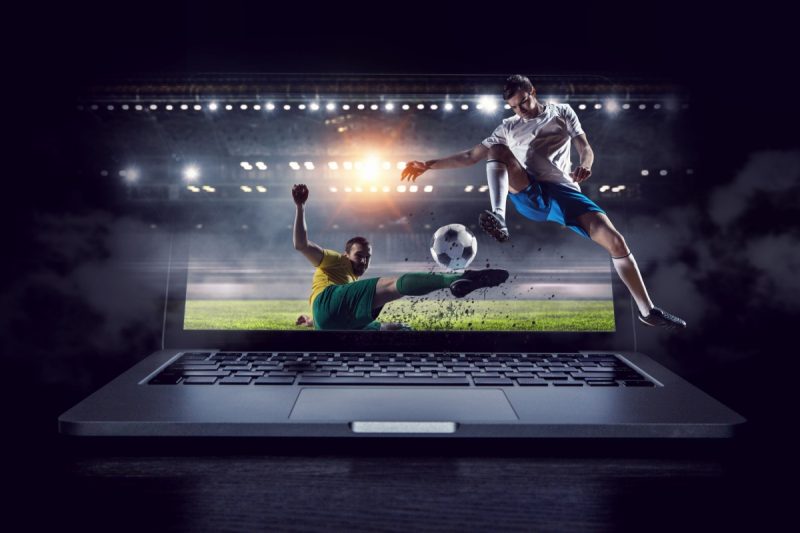 laptop care afiseaza doi jucatori de fotbal in timpul unui meci, cu tribuna unui stadion in fundal