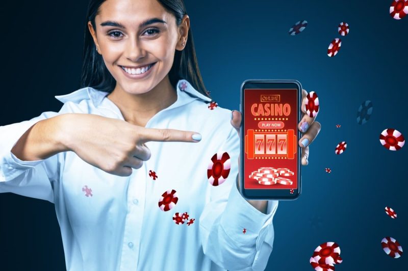 femeie zambitoare indicand spre un telefon mobil pe care ruleaza o aplicatie de cazino online