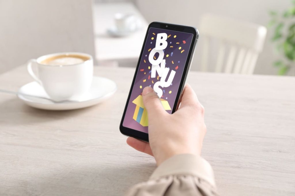persoana stand la masa, cu o ceasca de cafea, tinand in mana un telefon mobil pe care este afisata o notificare bonus