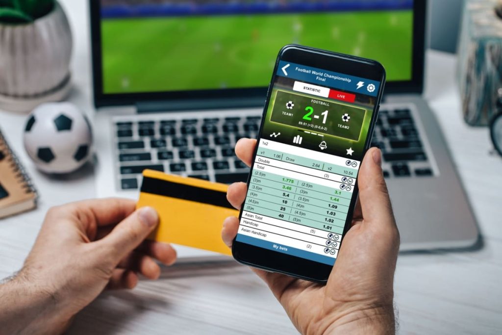 barbat in fata unui laptop, cu un card de credit intr-o mana si un telefon mobil pe care sunt afisate statisticile unui meci de fotbal in cealalta