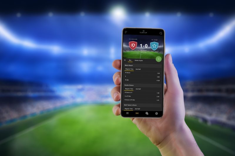 persoana tinand in mana un telefon mobil pe care este afisat scorul live al unui meci de fotbal, cu stadionul in fundal