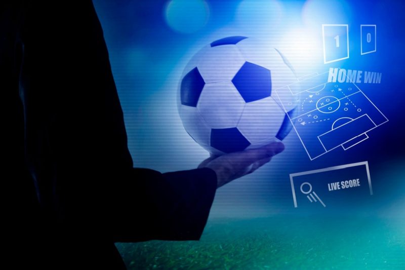 persoana tinand in mana o minge de fotbal, cu o tabela de scor si schema de joc a unei unui echipe in fundal