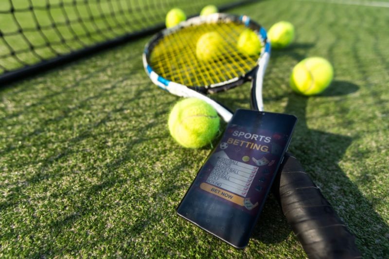 telefon mobil, afisand o aplicatie de pariuri sportive, alaturi de o racheta si mingi de tenis pe un teren