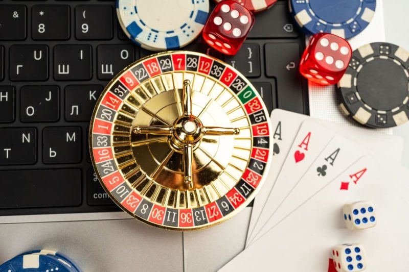 mini-ruleta, zaruri, carti de joc si jetoane de cazino, alaturi de un laptop