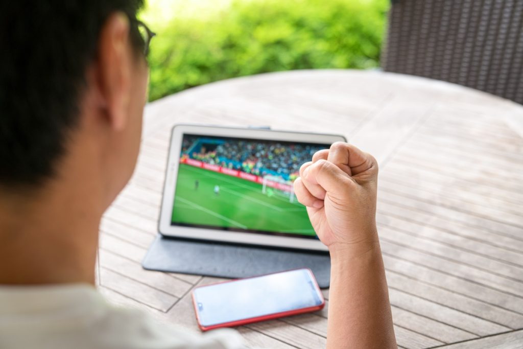 barbat asezat la o masa, cu o tableta in fata, pe care urmareste un meci de fotbal