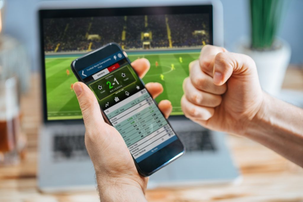 barbat sarbatorind rezultatul unui meci de fotbal, cu telefonul in mana pe care este afisat scorul meciului si un laptop in fundal