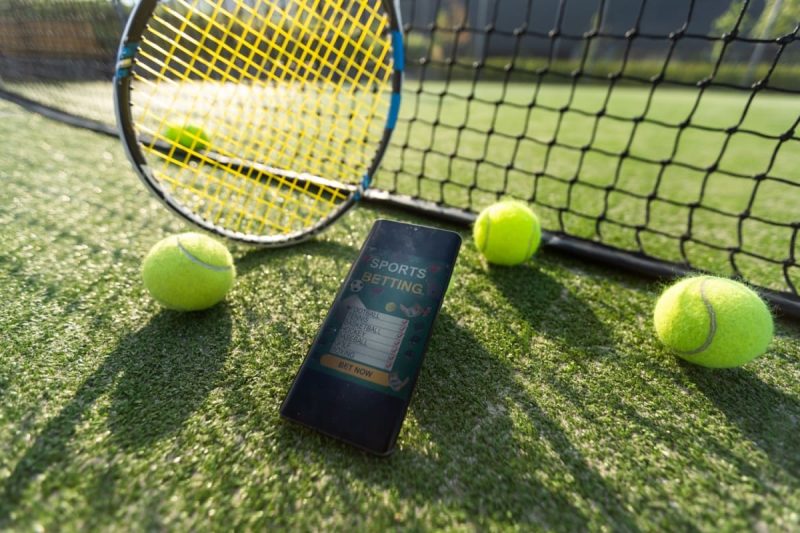 telefon mobil, mingi de tenis si racheta sprijinite de fileu pe un teren de tenis cu iarba
