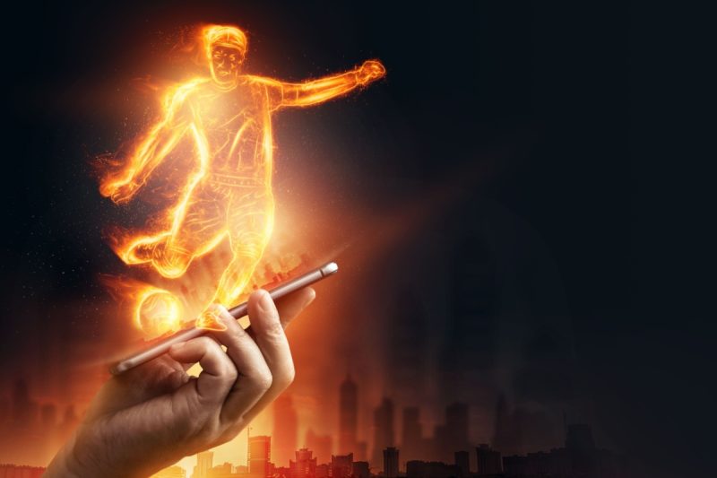 mana tinand un telefon mobil din care se proiecteaza o holograma a unui jucator de fotbal