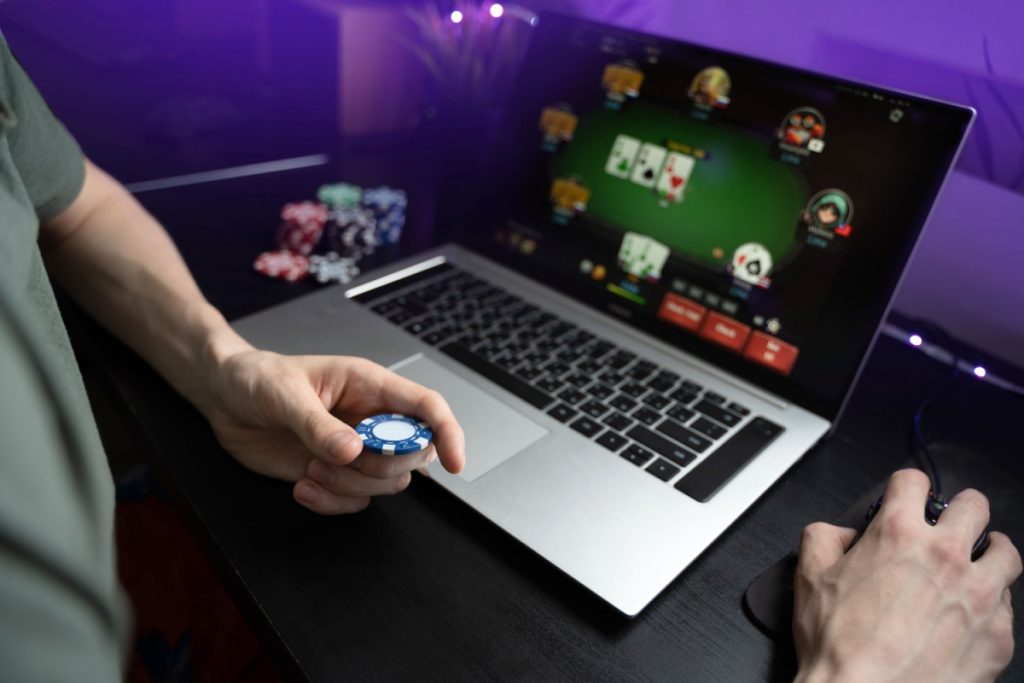 barbat in fata unui laptop pe care ruleaza un joc de poker, cu un jeton de cazino in mana