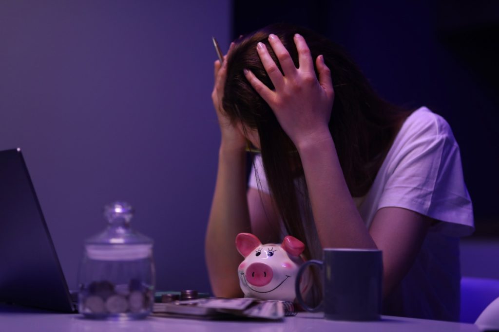 femeie trista in fata unui laptop, cu pusculite si caiet cu cheltuilelile lunare alaturi
