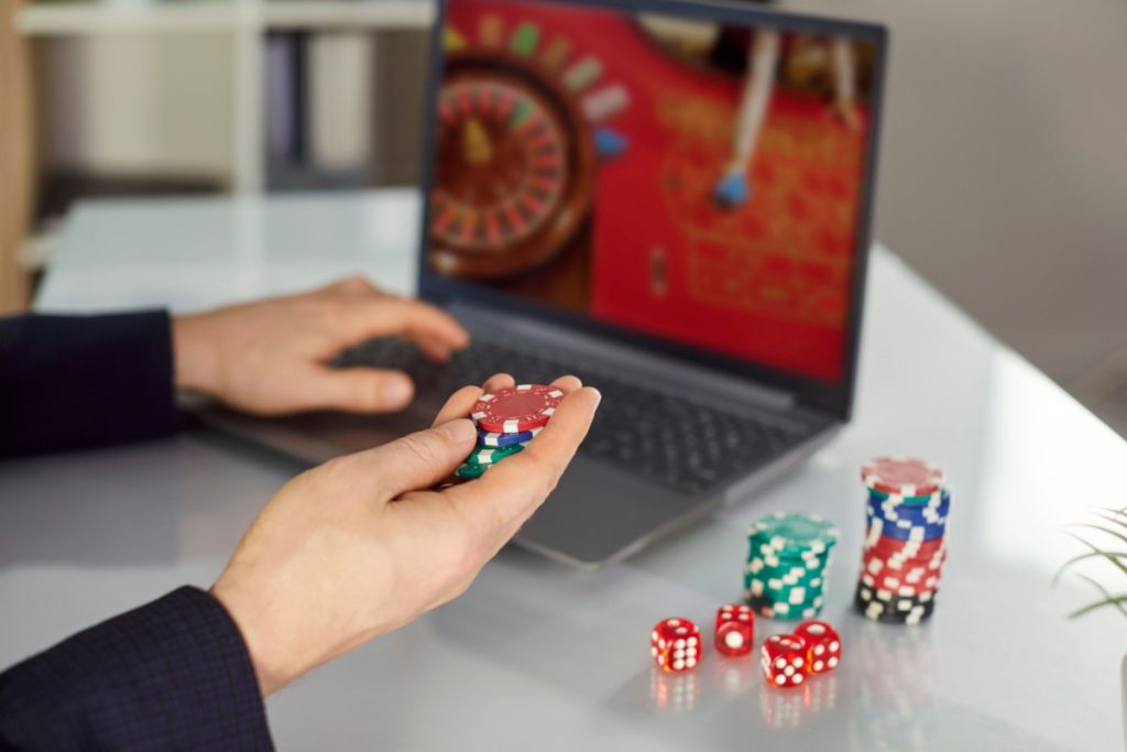 barbat in fata unui laptop pe care ruleaza un joc de ruleta online live, cu zaruri si jetoane de cazino alaturi