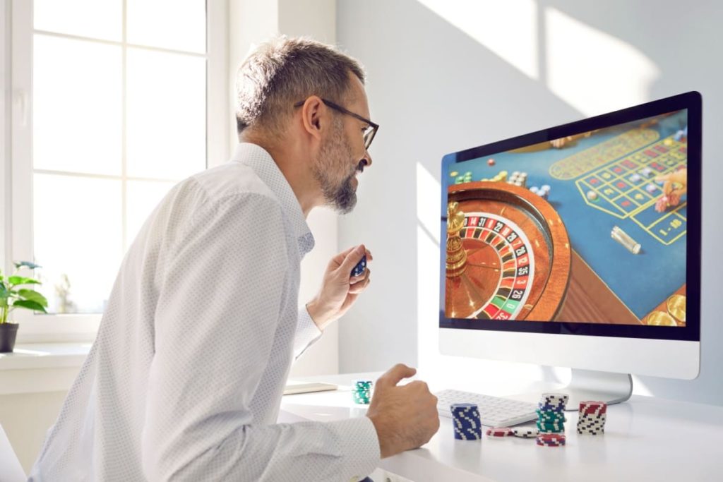 barbat entuziasmat urmarind un joc de ruleta live pe ecranul computerului, tinand in mana jetoane de cazino