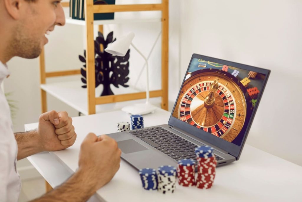 barbat fericit urmarind un joc de ruleta live cazino pe laptop, cu jetoane pe birou